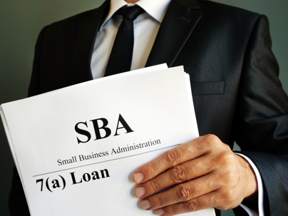 man in black and white suit and tie holding sheet of paper that says SBA Small Business Administration 7(a) loan
