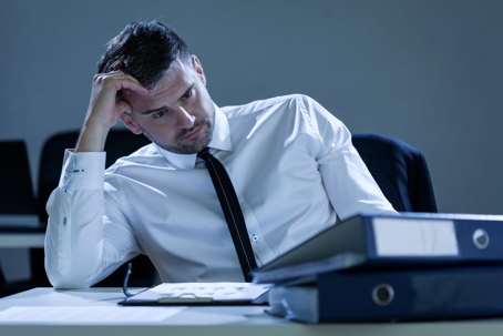 man in dress clothes with hand on head sitting at a desk with papers on the desk